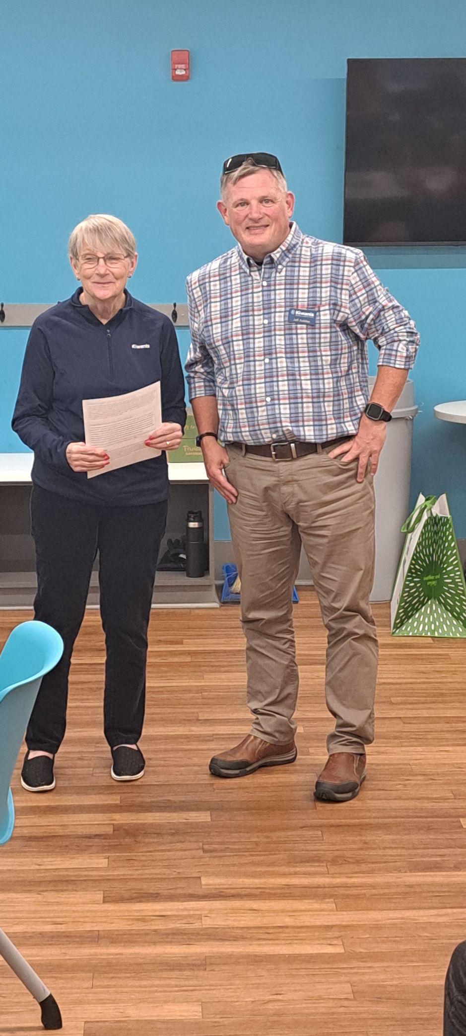 Two people stand indoors. A woman in dark clothes holds paper; a man wears a plaid shirt and tan pants.