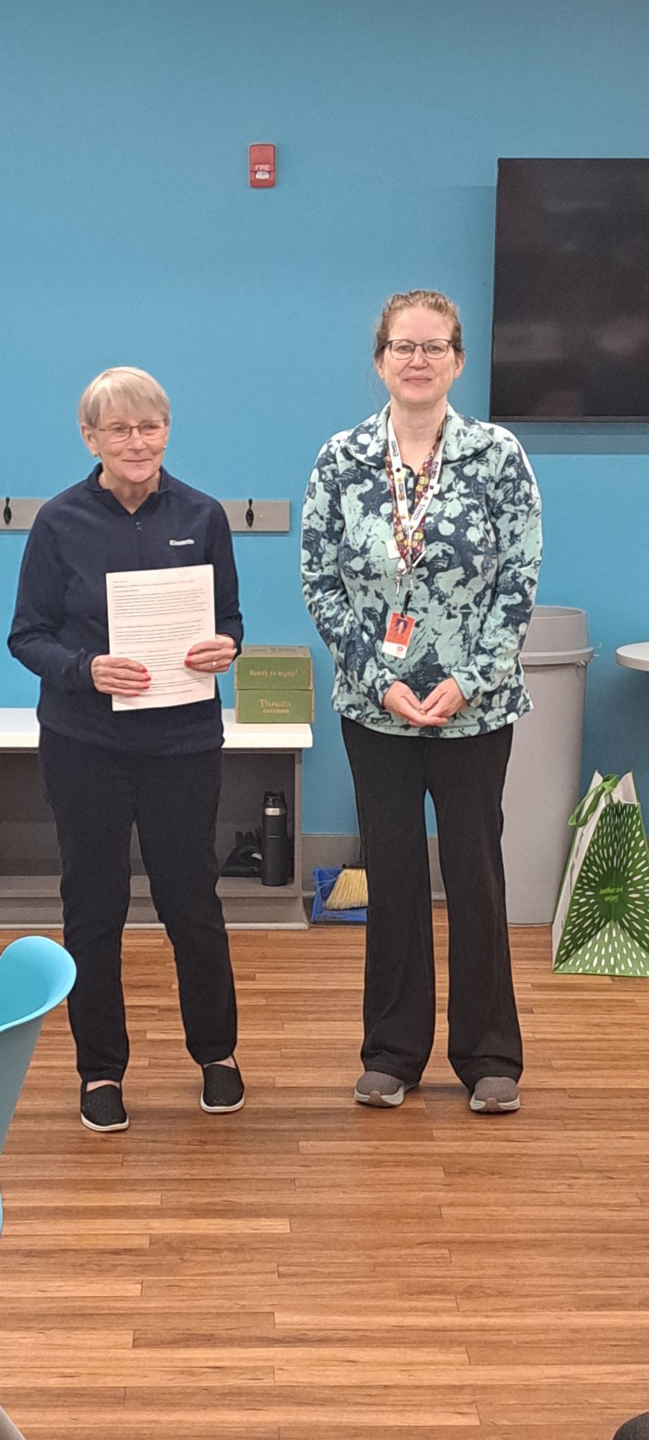 Two women stand indoors; one holds a paper, both wear dark pants. Blue wall in the background.