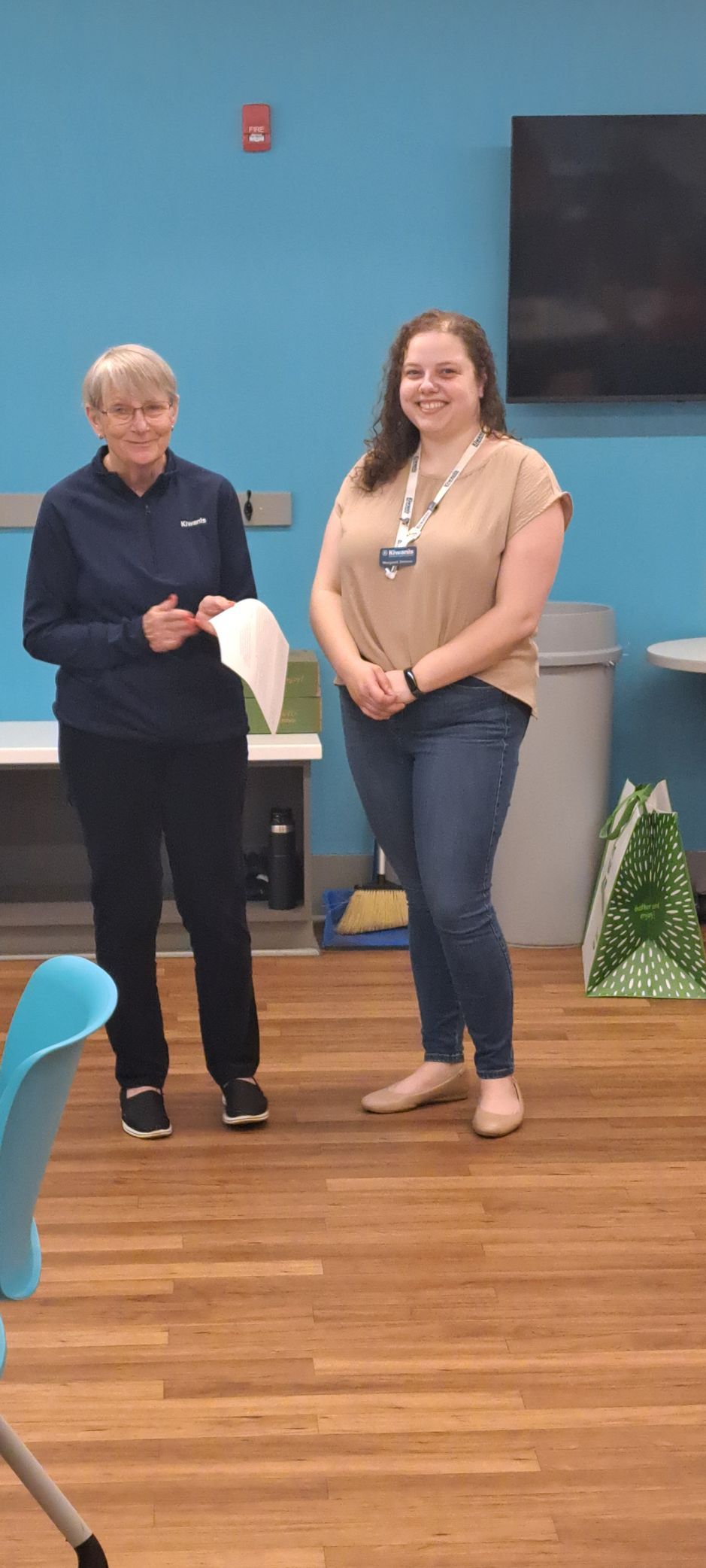 Two women standing in a room with blue walls and wooden floors. One holds papers and the other smiles.