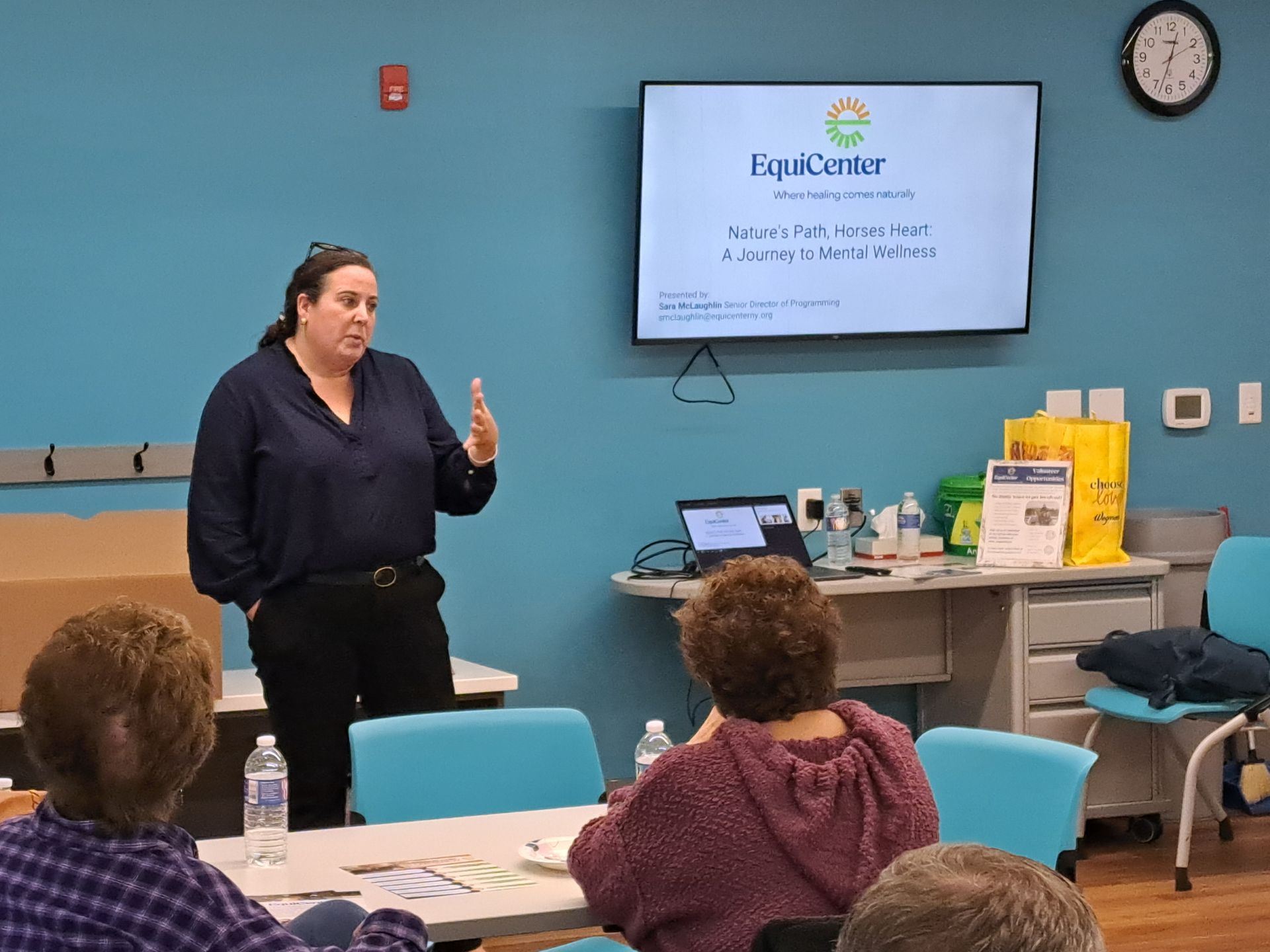 Woman giving a presentation in a room with a screen, seated audience, and table with water bottles.