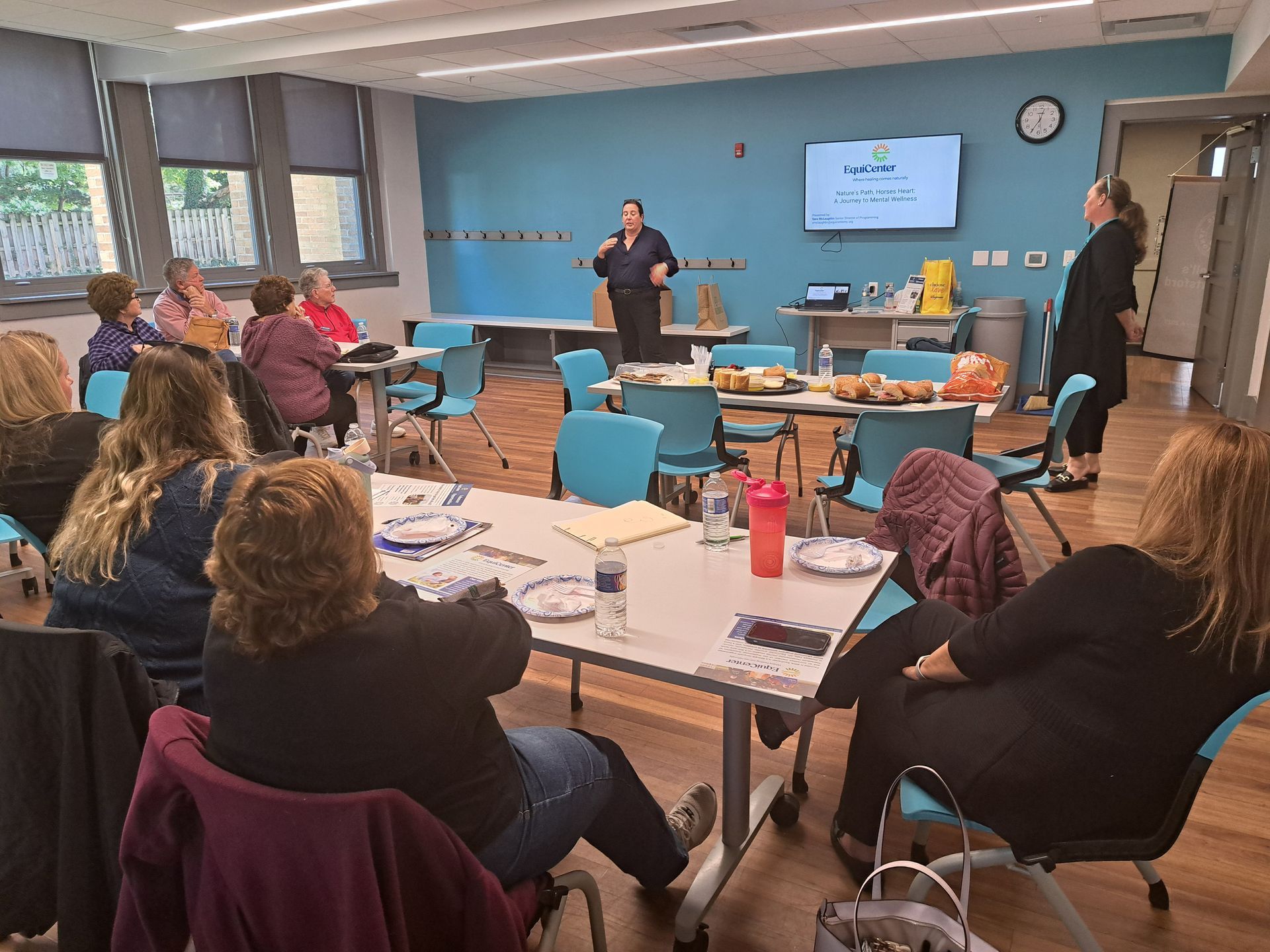 A presentation in a classroom: two women, audience of seated people, food on a table, a screen with text.