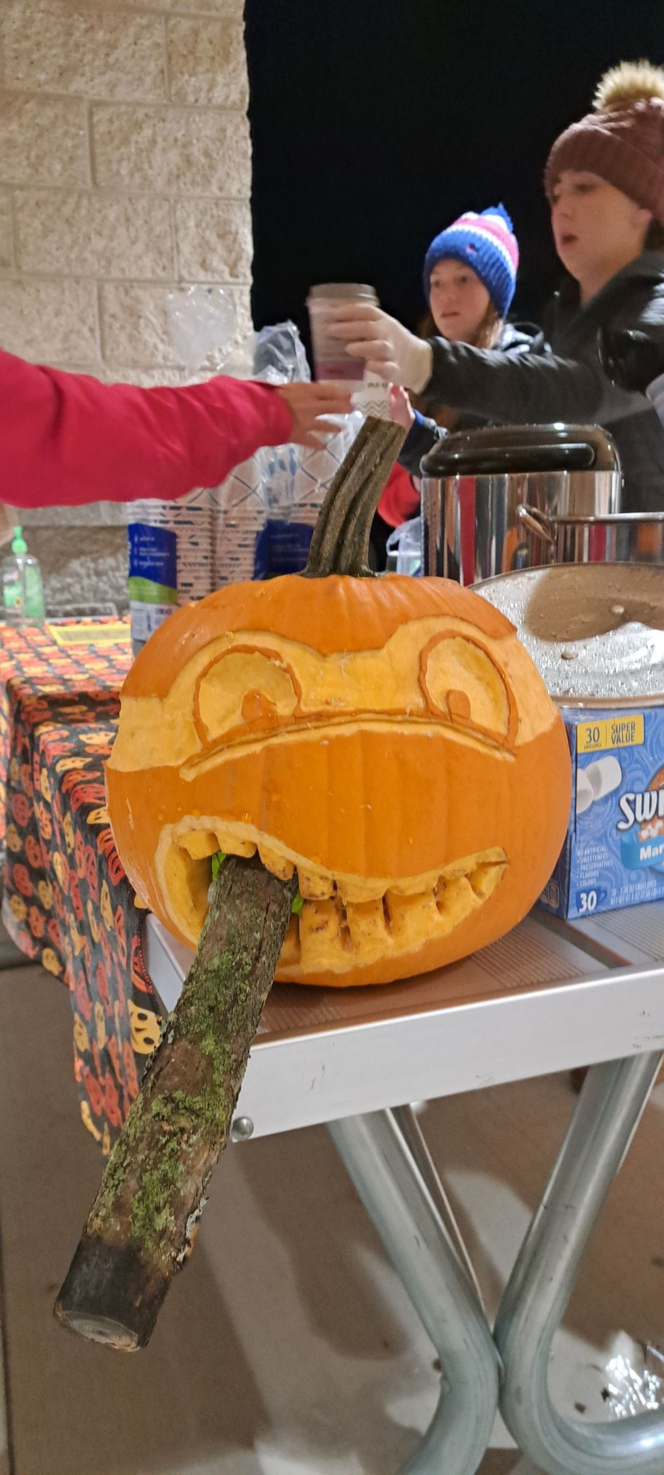 Carved pumpkin with a stick in its mouth; people in the background by a table.