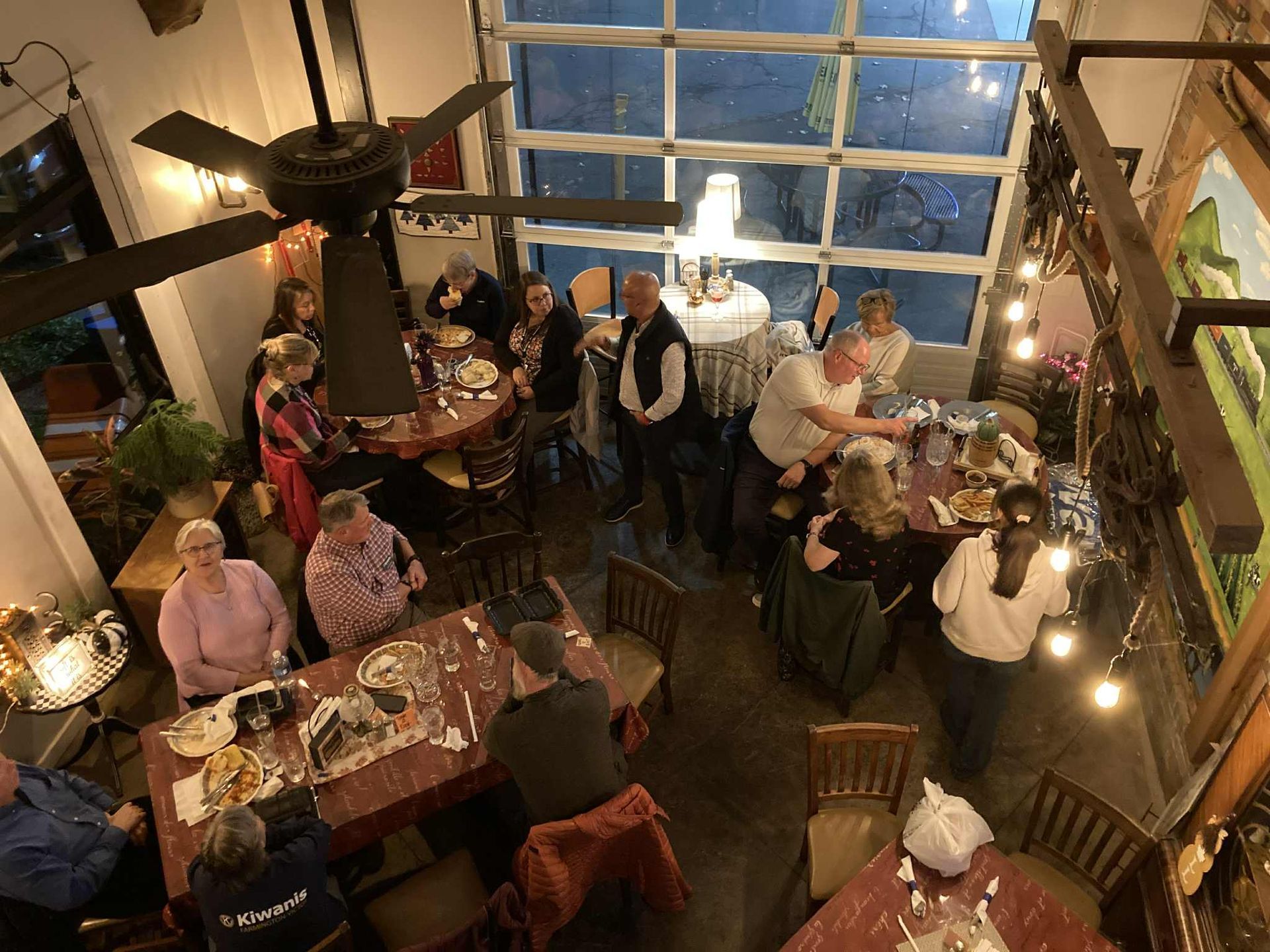People gather inside a restaurant with round tables, a glass garage door, and a ceiling fan.