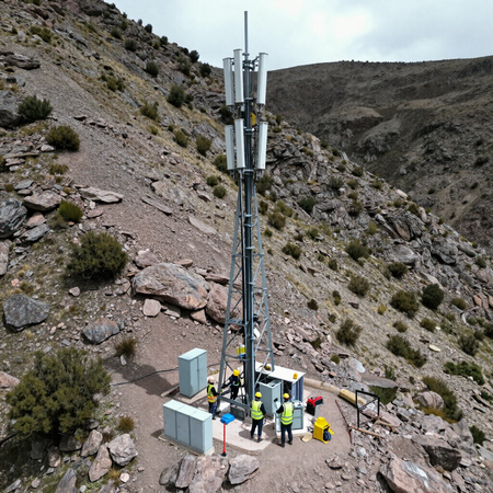 Unos operarios inspeccionan una alta torre de telefonía móvil situada en una ladera rocosa, con equipos en la base.
