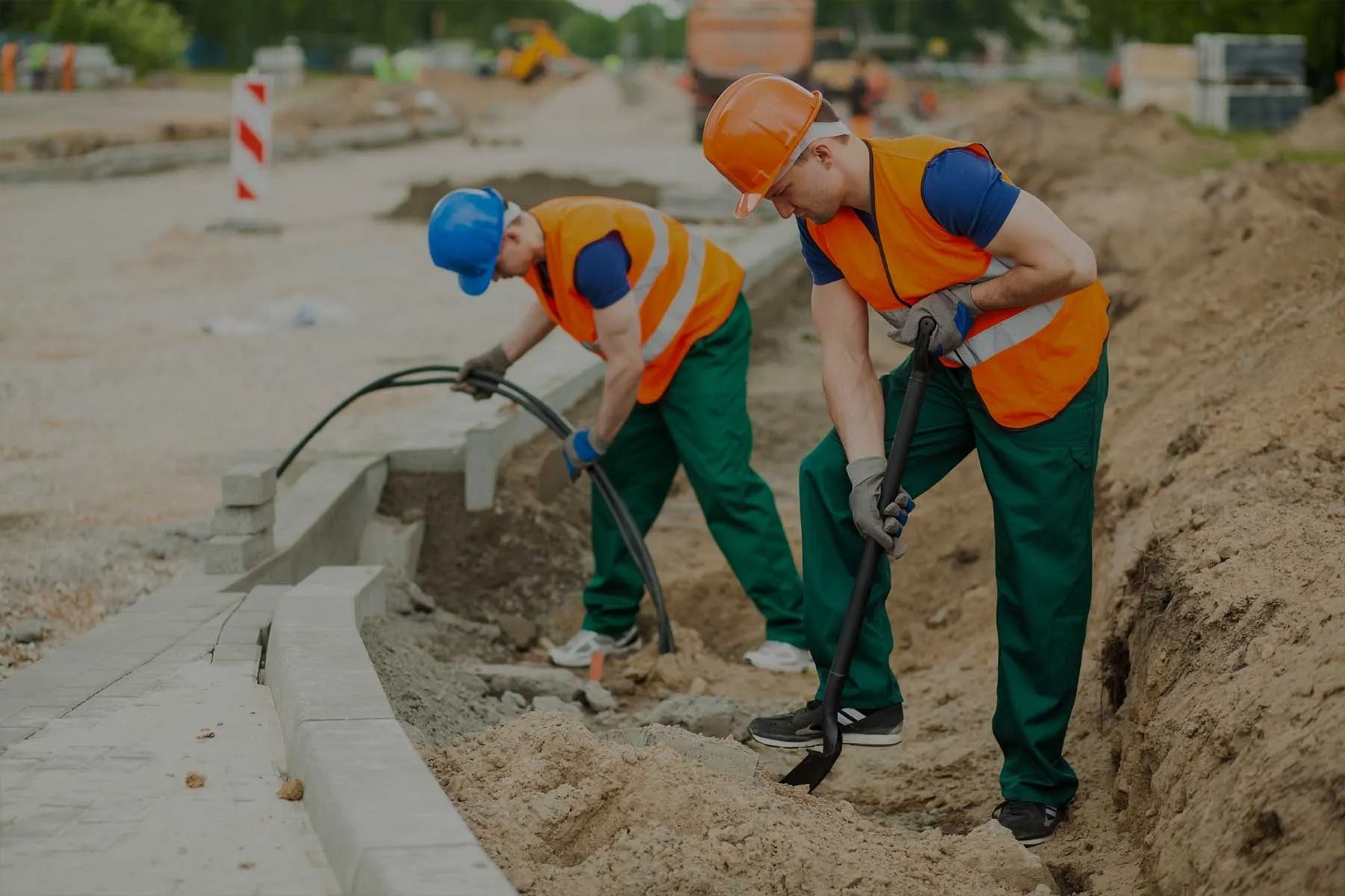 Tres trabajadores de la construcción con chalecos naranjas y cascos instalando un bordillo de hormigón en una obra de tierra.