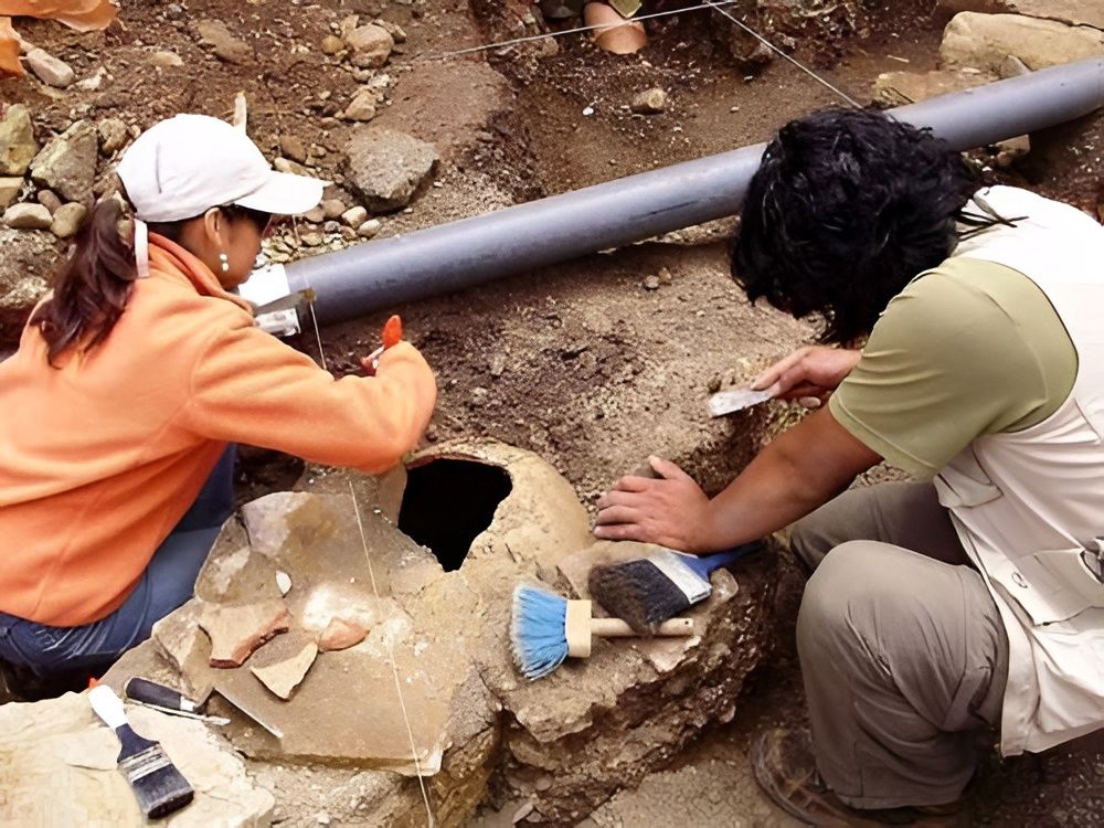 Dos trabajadores excavan alrededor de una tubería enterrada en una zanja rocosa con herramientas y una pala.
