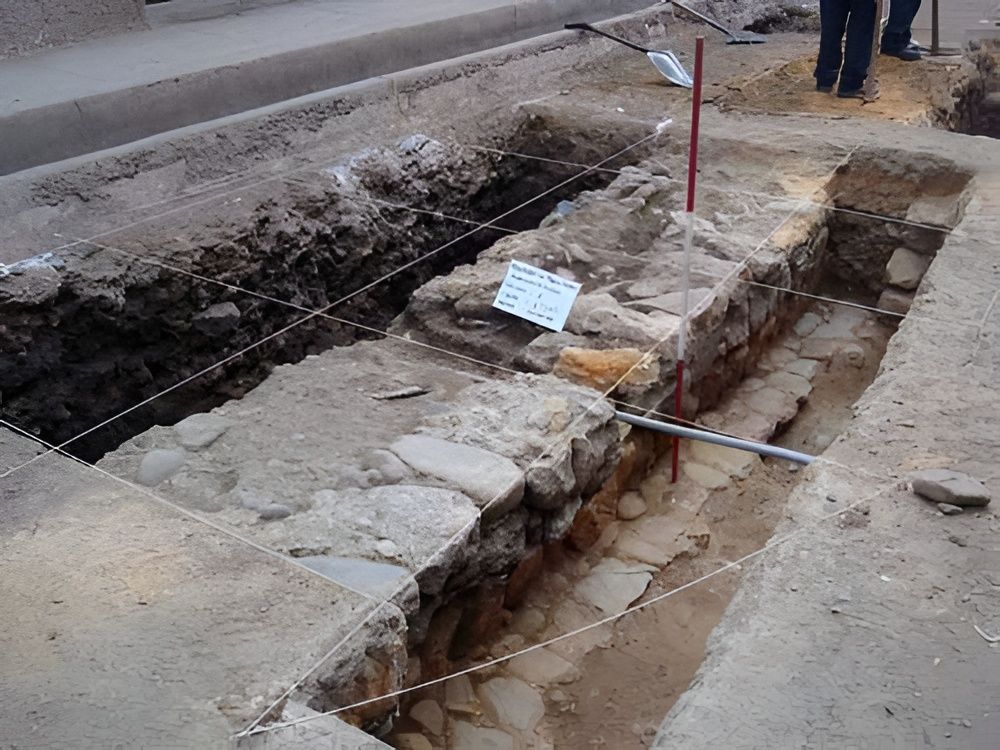 Excavación arqueológica con cimientos de piedra y una trinchera señalizada.