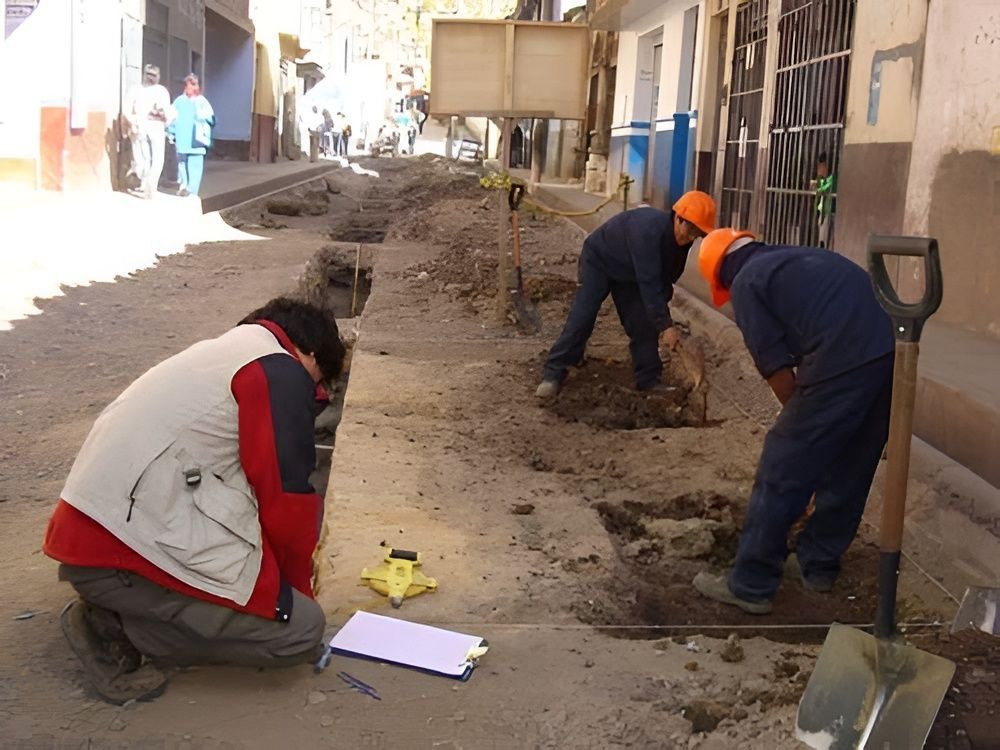 Obreros cavando una zanja en un callejón estrecho junto a un edificio, con herramientas y escombros en el suelo.