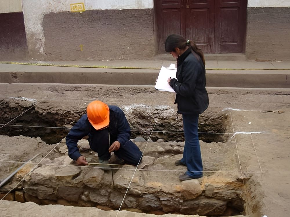 Obreros inspeccionando una zanja de piedra en una calle; uno lleva un casco naranja y el otro sostiene unos papeles.