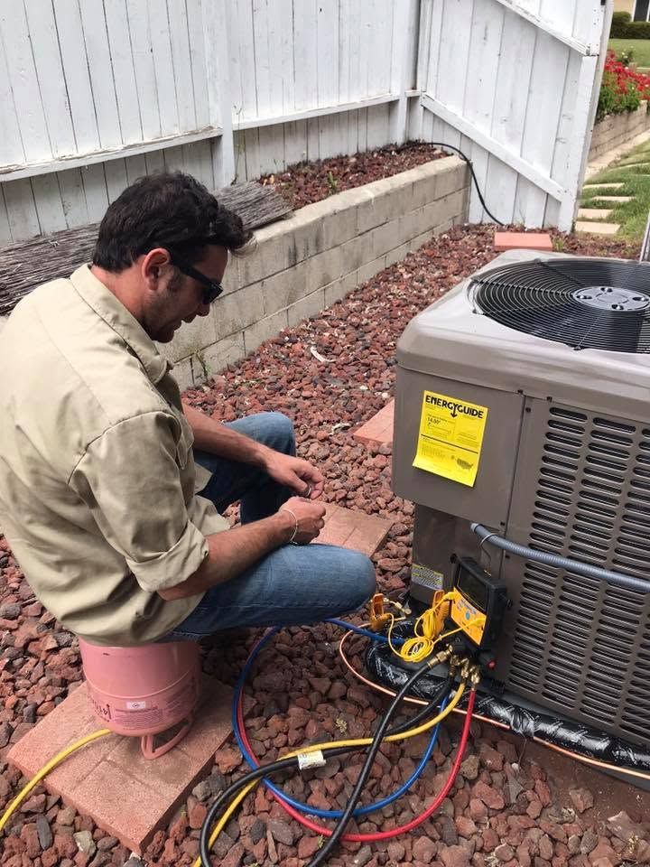 Technician kneeling beside outdoor air conditioner, working on wiring with tools and gauges.