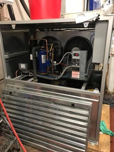 Open metal appliance with exposed compressor and wiring, front panel removed in a workshop.