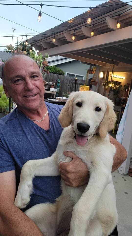 Man holding a cream-colored puppy outdoors under string lights, both facing the camera.