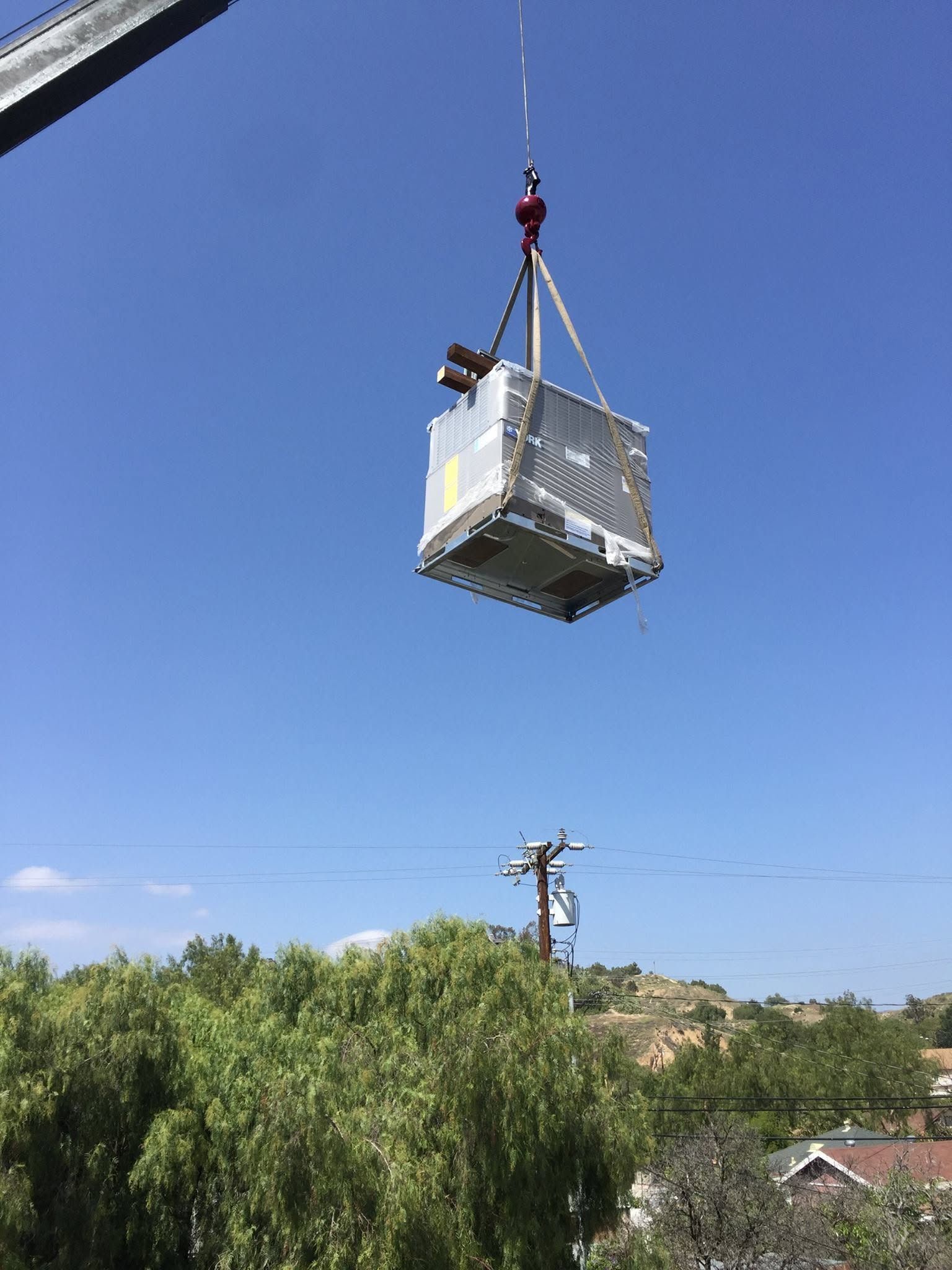 Crane lifting a wrapped metal container over trees under a clear blue sky