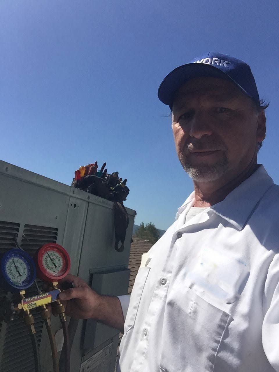 Man in white shirt and cap holding HVAC gauges beside an outdoor unit under a clear blue sky