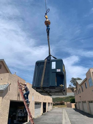 Crane lifting a large blue industrial unit over a warehouse alley beside a worker on a ladder