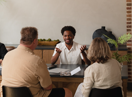 Three people seated at a table in a meeting, with one person speaking and gesturing in a bright room.