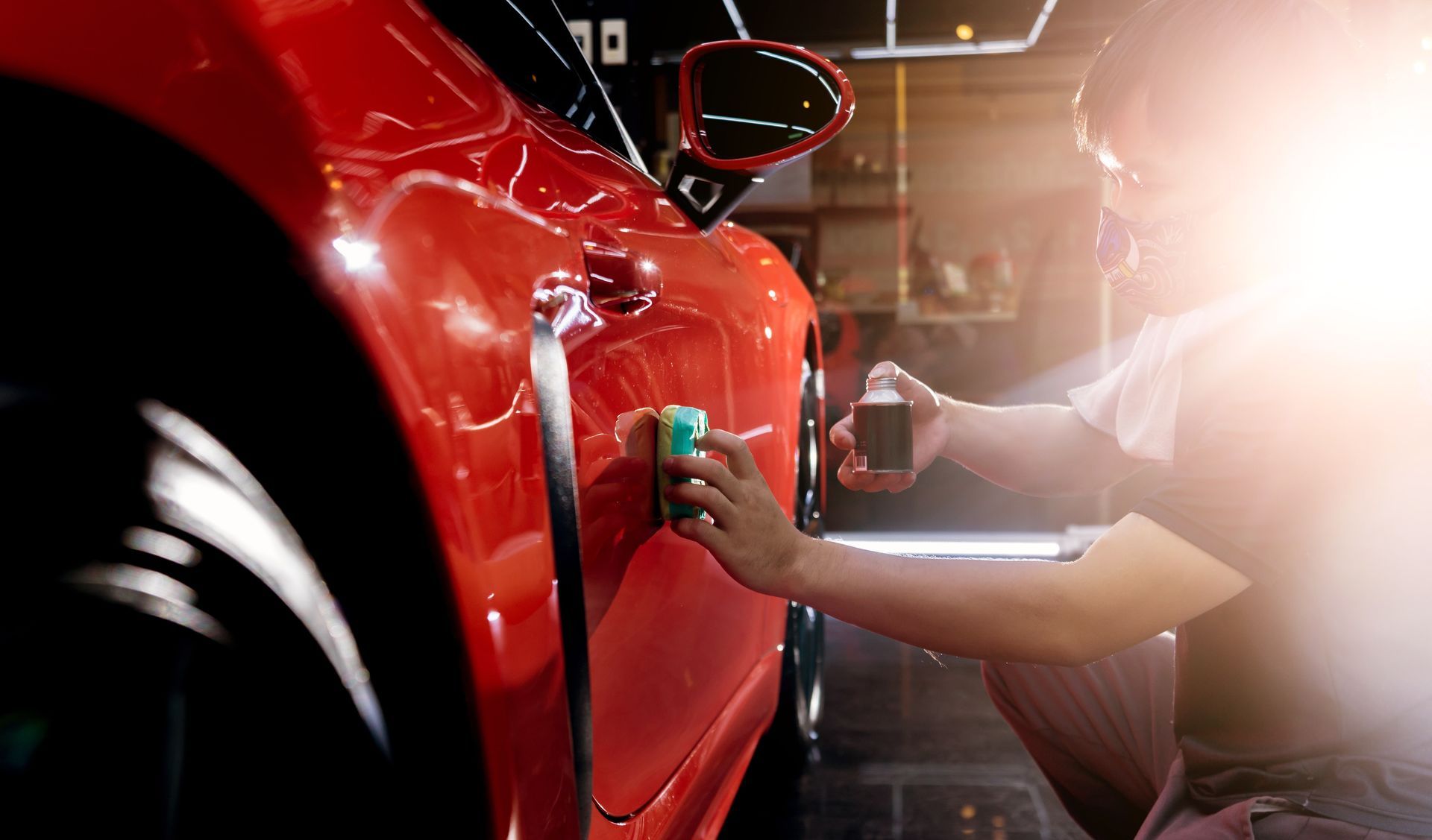Person polishing a red car door in a garage with bright light glare