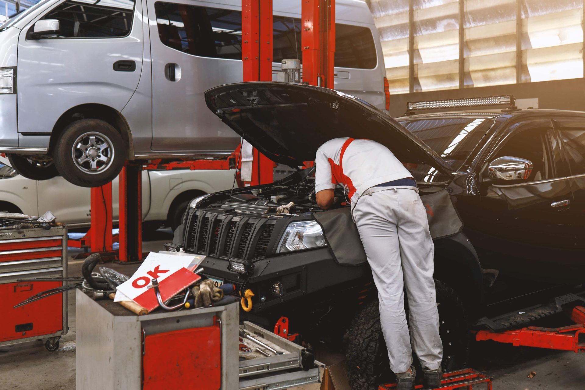 Mechanic working on a black car in a garage with another car lifted on a lift in the background
