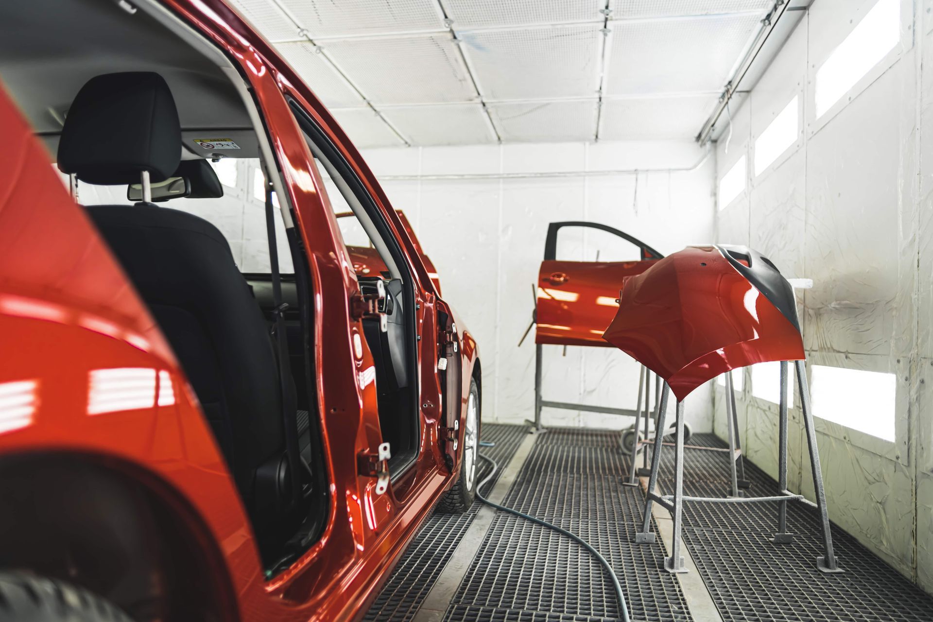 Red car body in an auto paint booth with a detached hood on a stand and another car door in the background
