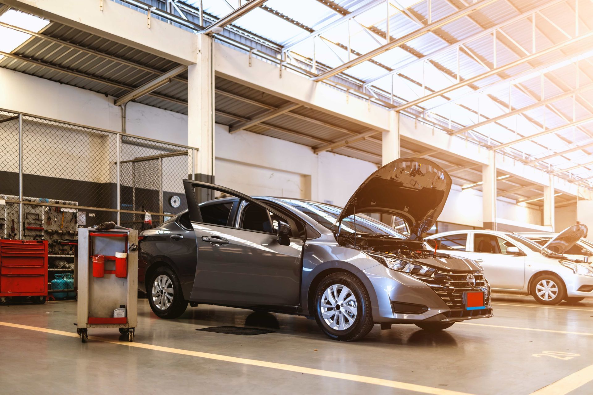 Cars in a bright auto repair garage, one with its hood open and a mechanic’s cart nearby