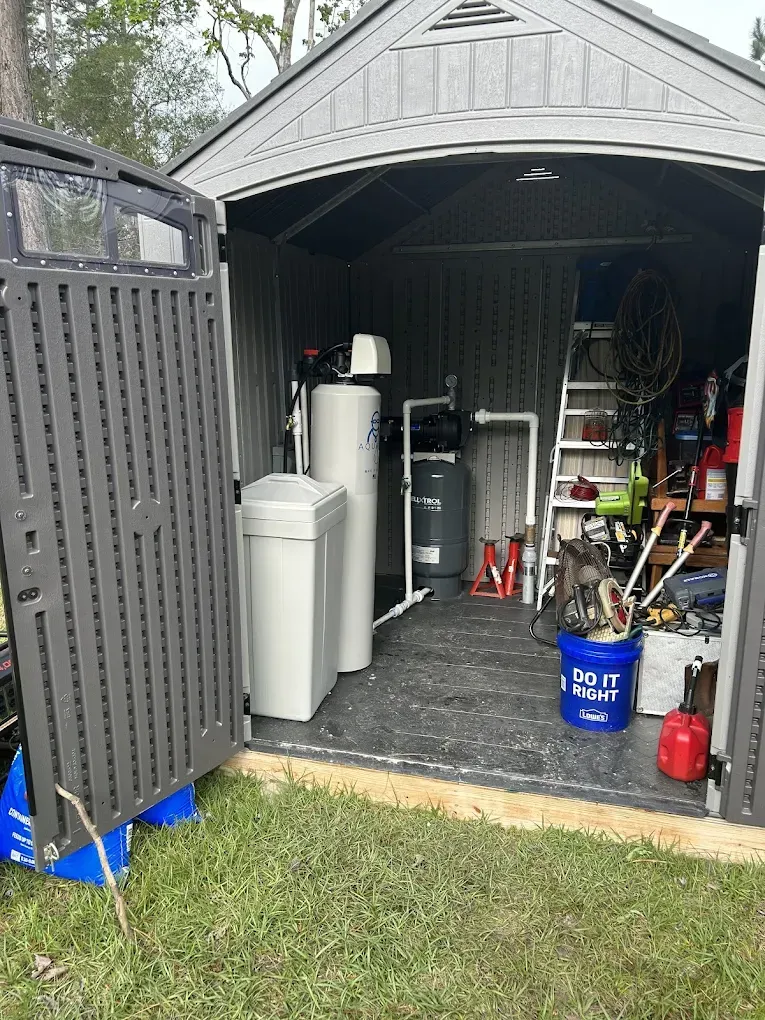 Gray shed interior with water treatment system, tools, and a ladder. The door is open, revealing the contents.