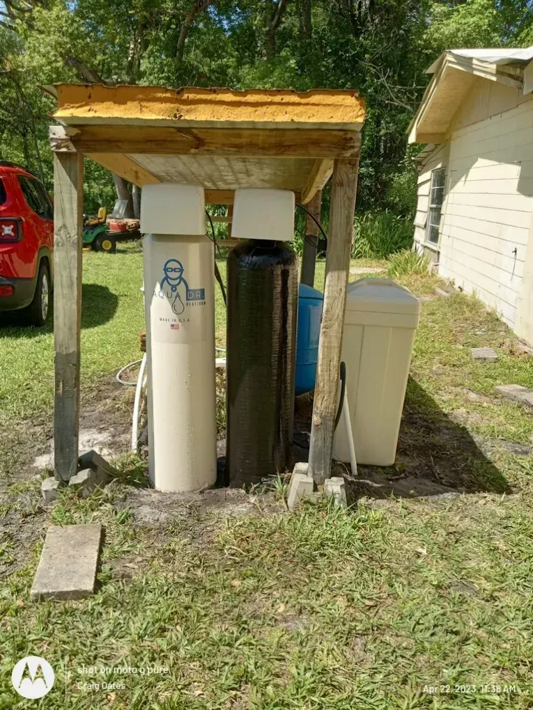 Outdoor water filtration system under a wooden shelter, tan and black tanks, white container.