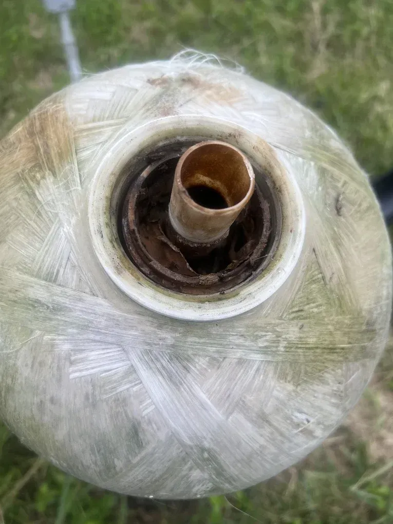 Top-down view of a damaged water tank, brown pipe and corroded opening in the center. Light-colored, fibrous tank exterior.