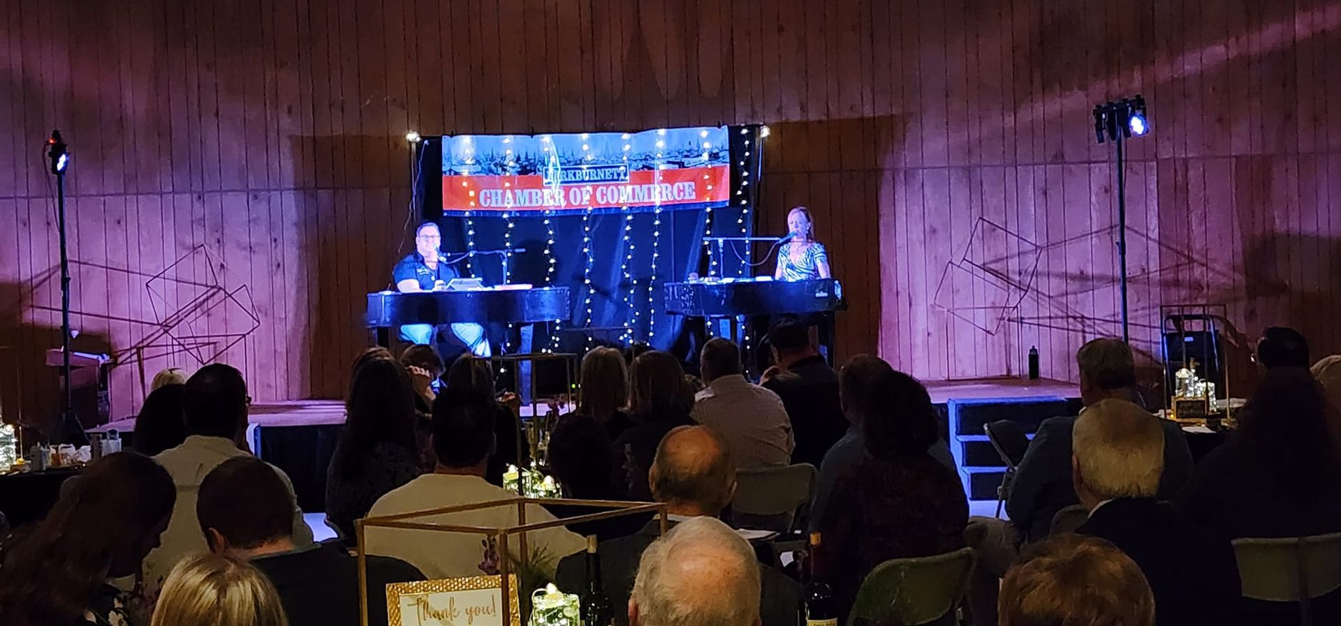 Two people playing pianos on a stage in front of an audience in a dim room with purple lighting.