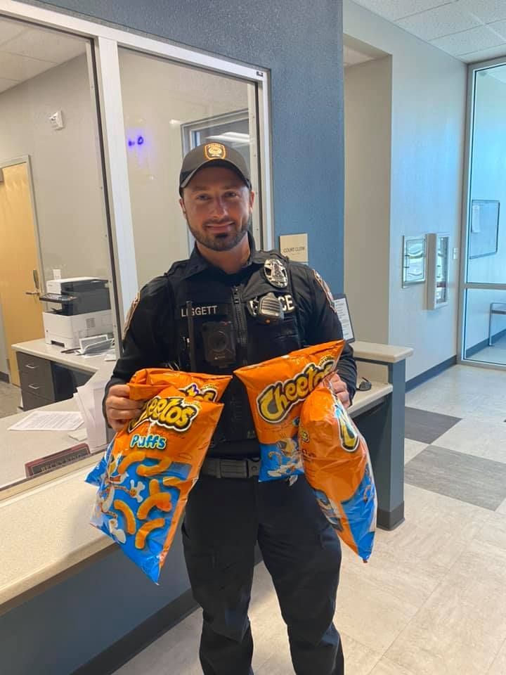 Police officer holding two large bags of Cheetos in an office setting.