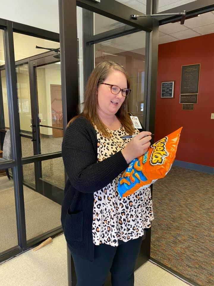 Woman smiling, holding open bag of Cheetos, standing inside a building.