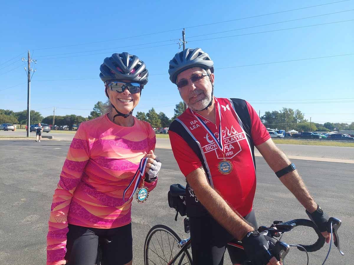 Cyclists wearing helmets and medals pose with a bicycle in a parking lot. Woman in pink shirt smiles. Man in red shirt. Sunny day.