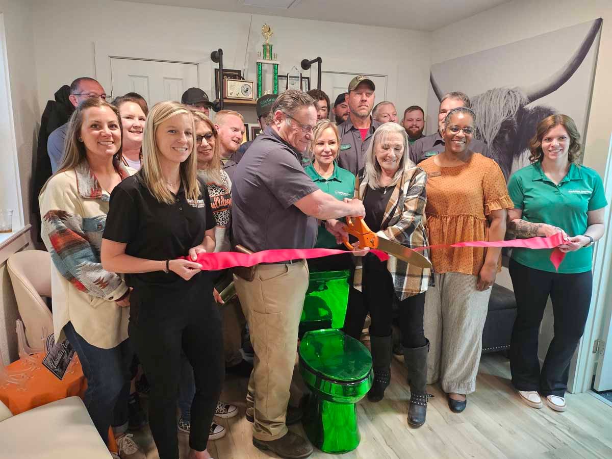 Group cuts a red ribbon at a business opening. People watch, some smiling, in a room.