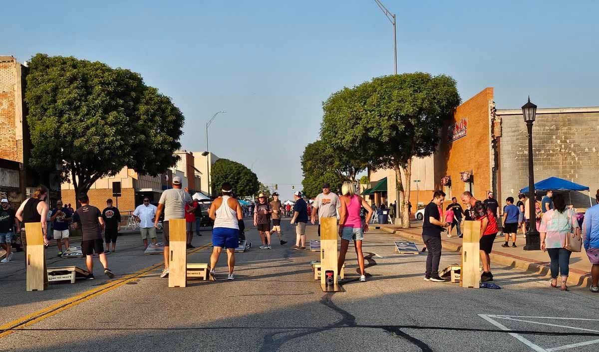 People in a street with wooden platforms. Buildings line the sides; trees and blue sky.