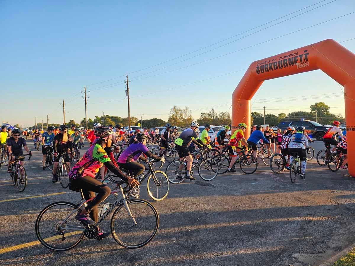 Cyclists start a race under an orange inflatable archway on a paved road.