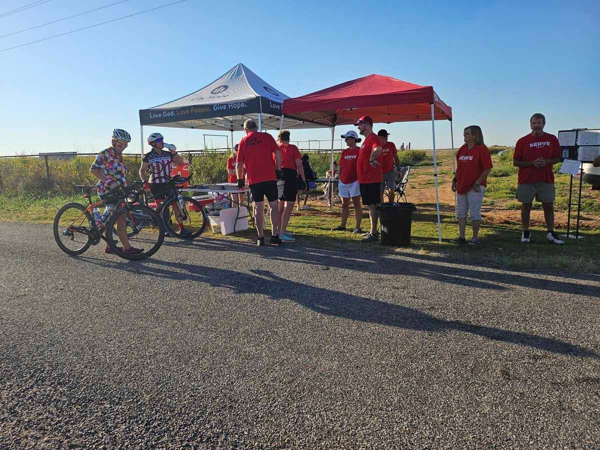 Cyclists at rest stop, wearing red shirts, with canopy tents, and bicycles on gravel road.
