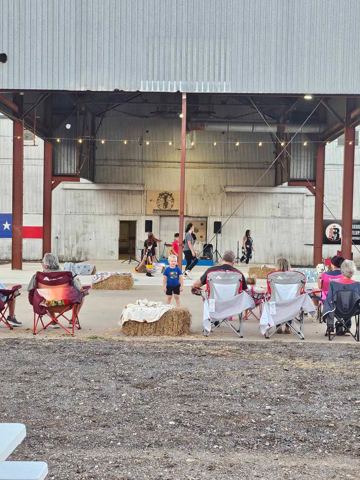 Outdoor event: People seated in chairs watch a performance on a stage, string lights overhead.