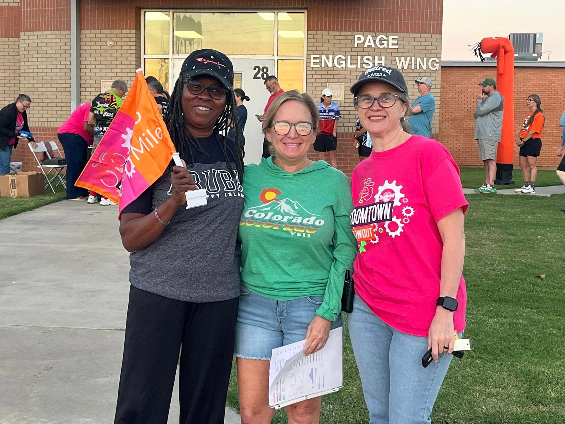 Three women pose in front of a building. One holds a flag, another a paper, and the third wears a pink shirt.