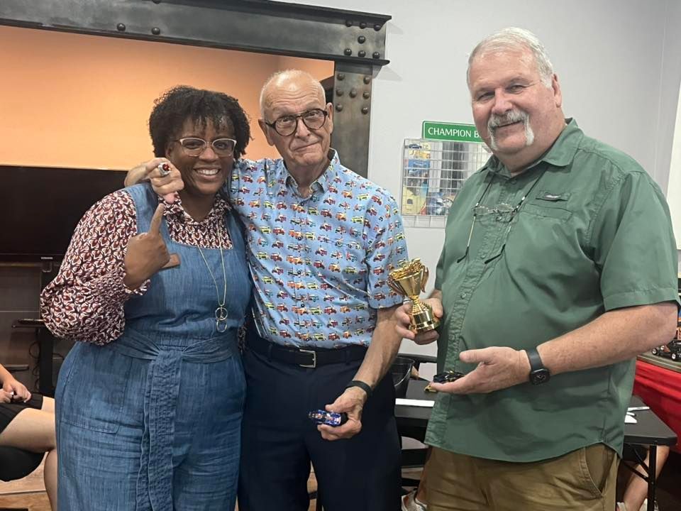 Three people smiling, posing with a trophy indoors. Woman in denim jumpsuit gives thumbs up, man holds trophy, another man embraces them.