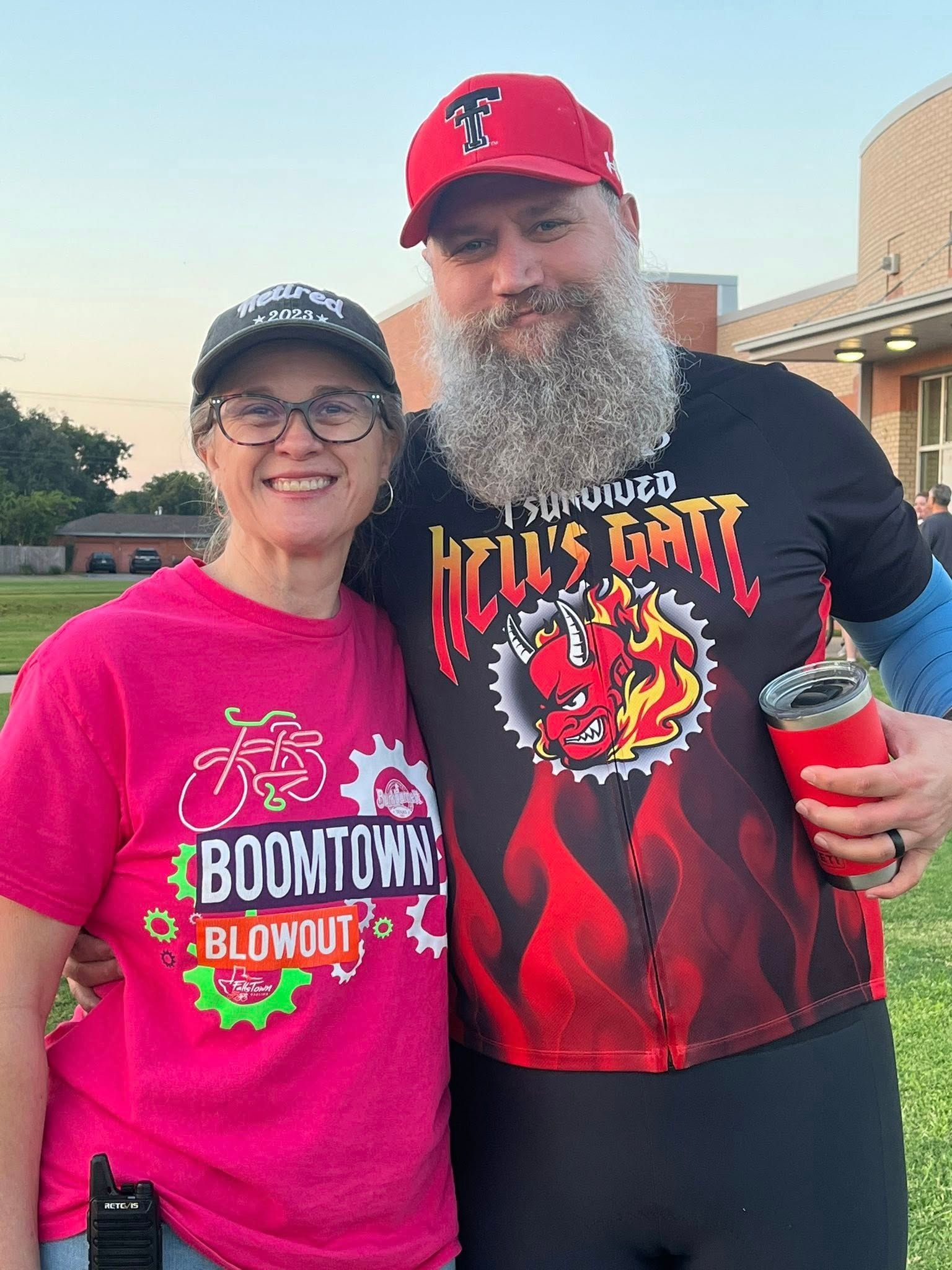 Woman in pink shirt and man with beard pose outdoors. Man holds red cup. Buildings in background.