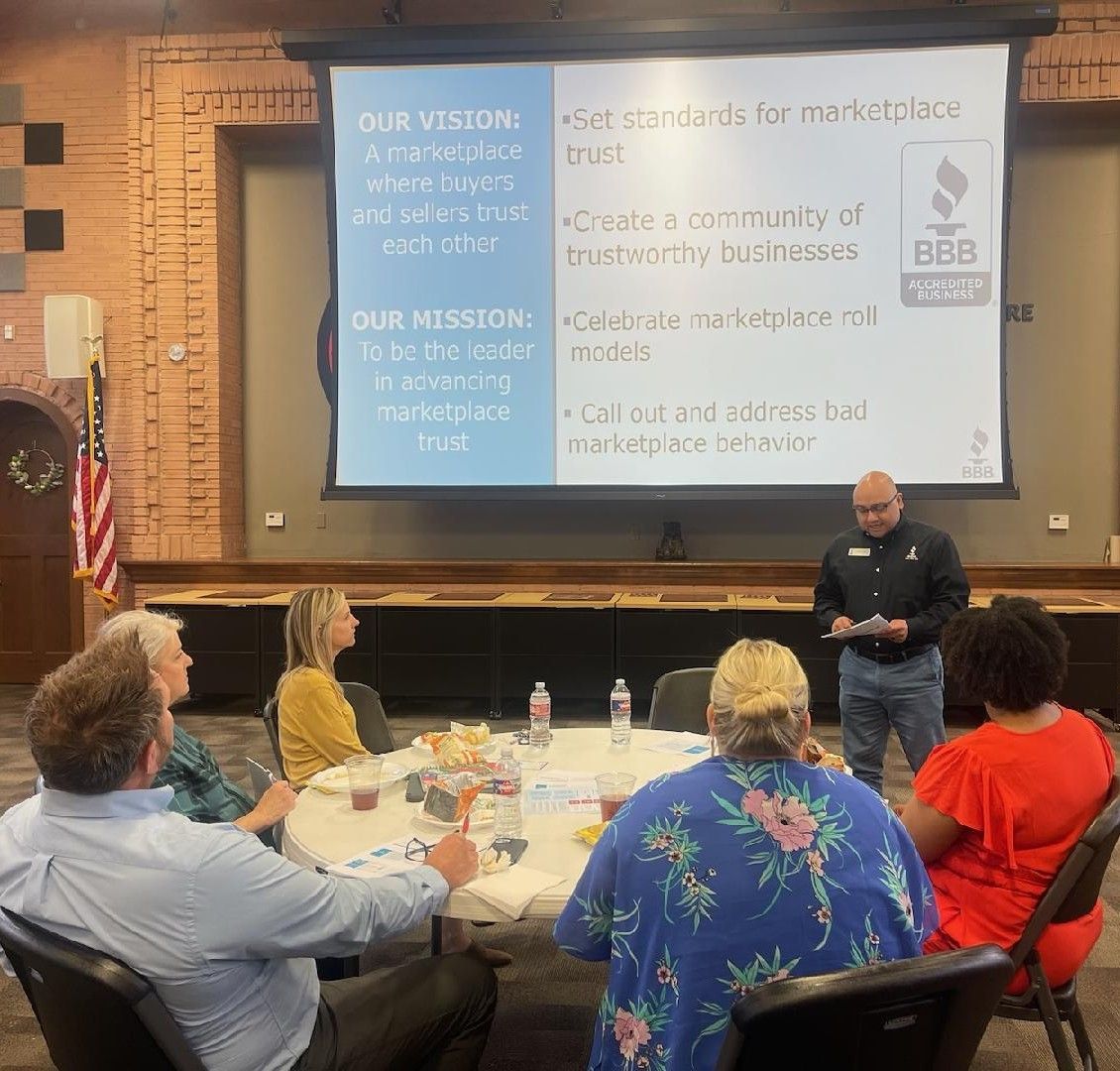 Man presenting to a group seated around a table; a screen displays a vision statement.
