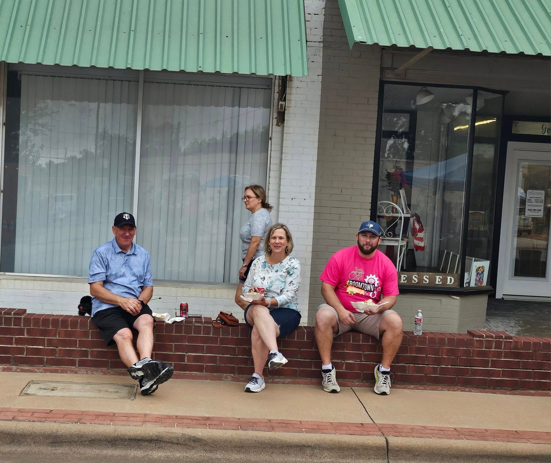 People sitting on a brick wall in front of storefronts, under green awnings.