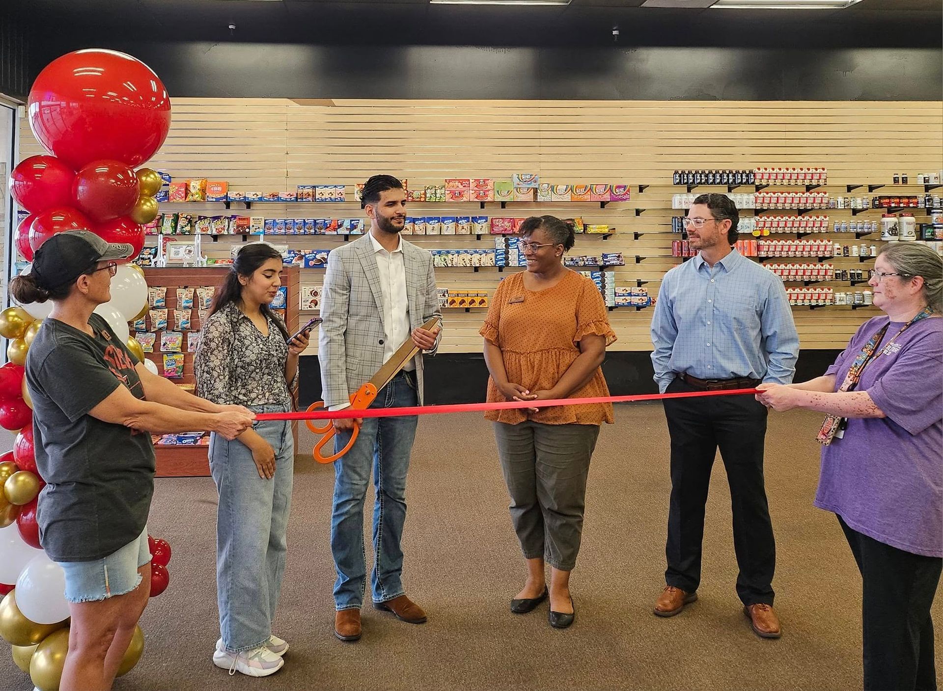 People cutting a ribbon at a store opening; red balloon arch; shelves of merchandise in the background.