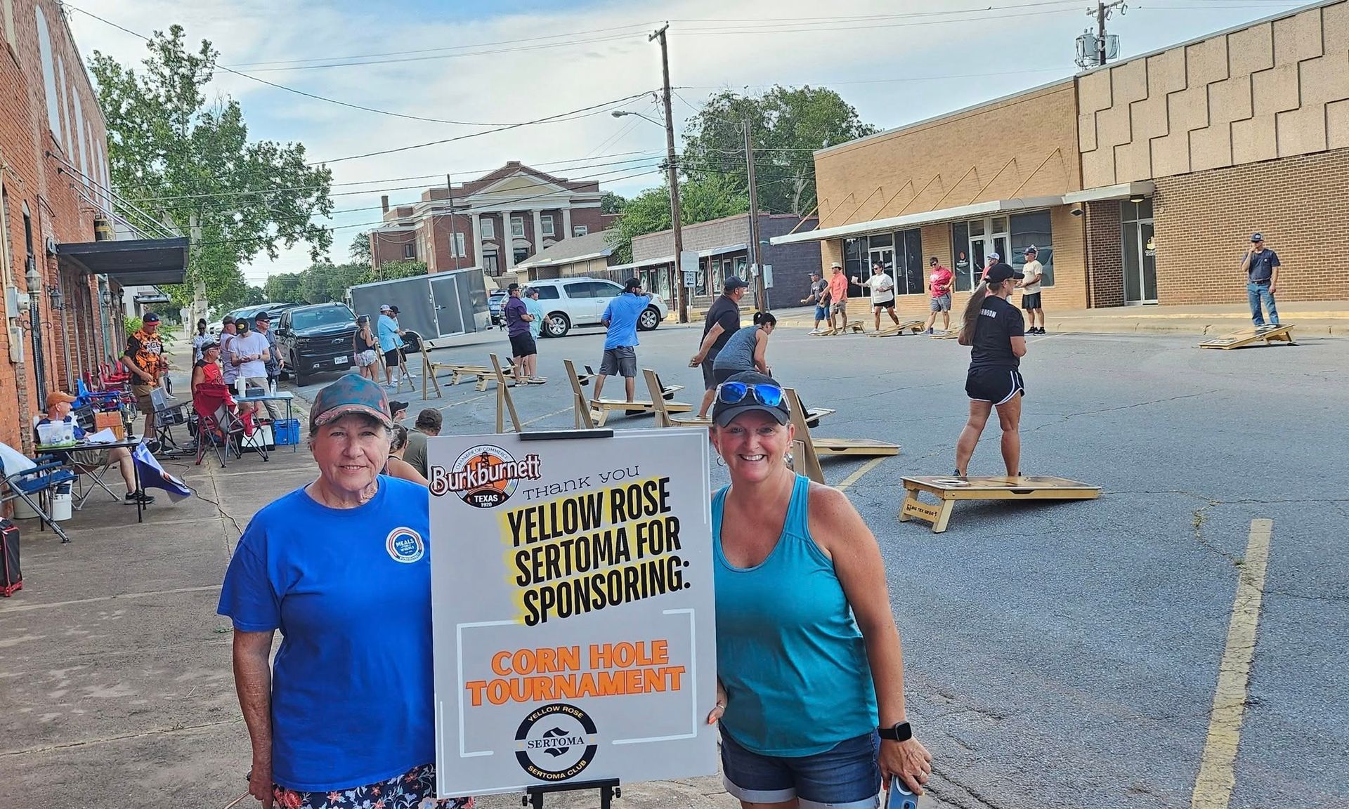 Two women by a sign for a cornhole tournament. People play in a street lined with buildings.