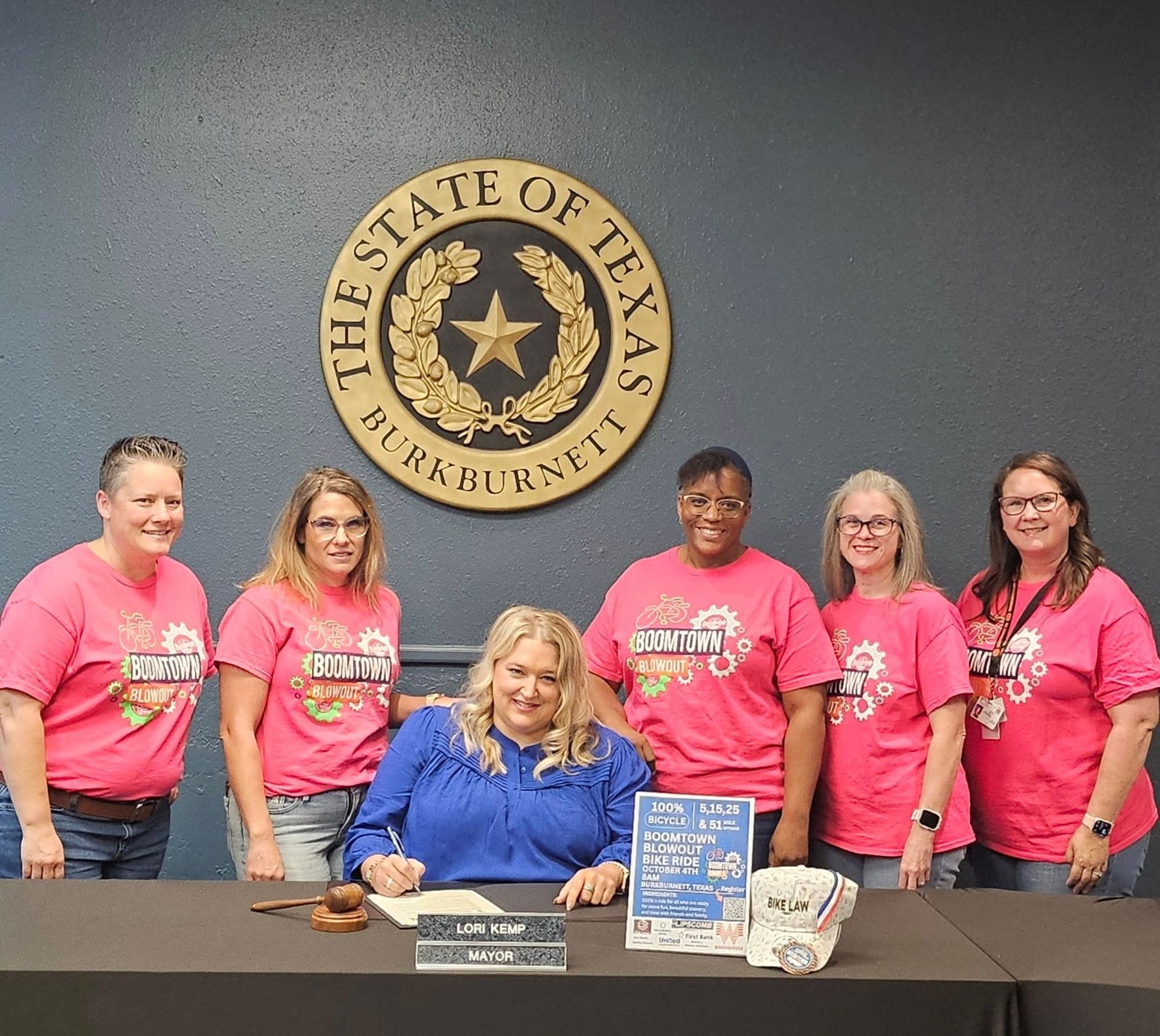 Group of people in pink shirts and blue shirt around a table, signing a document in front of a Texas seal.