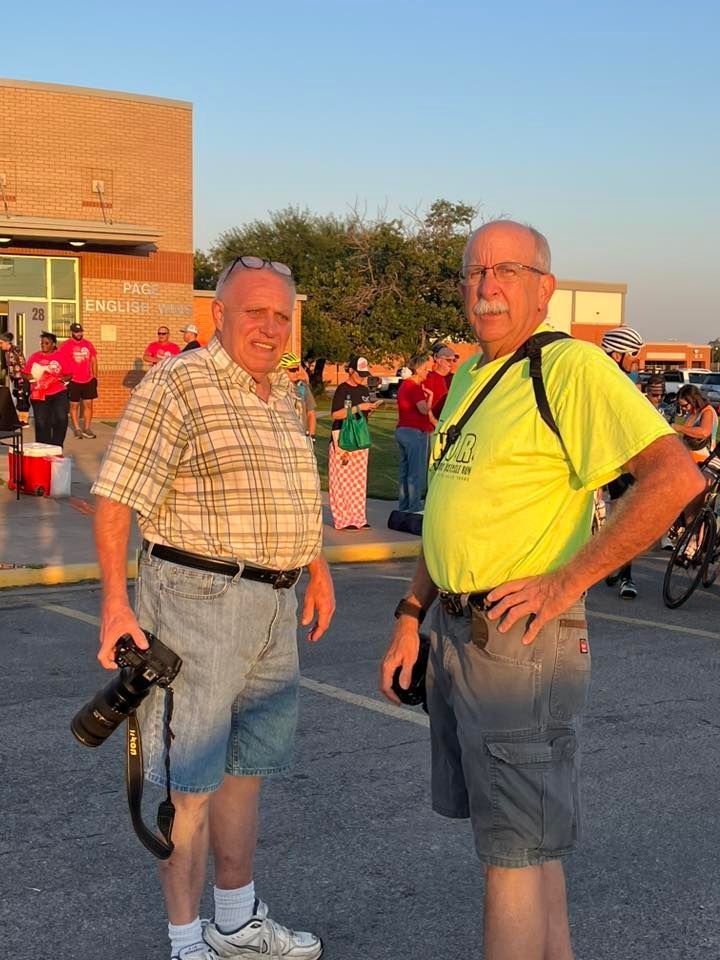 Two older men standing in front of a building; one holding a camera, one wearing a yellow shirt and shorts; outdoor setting.