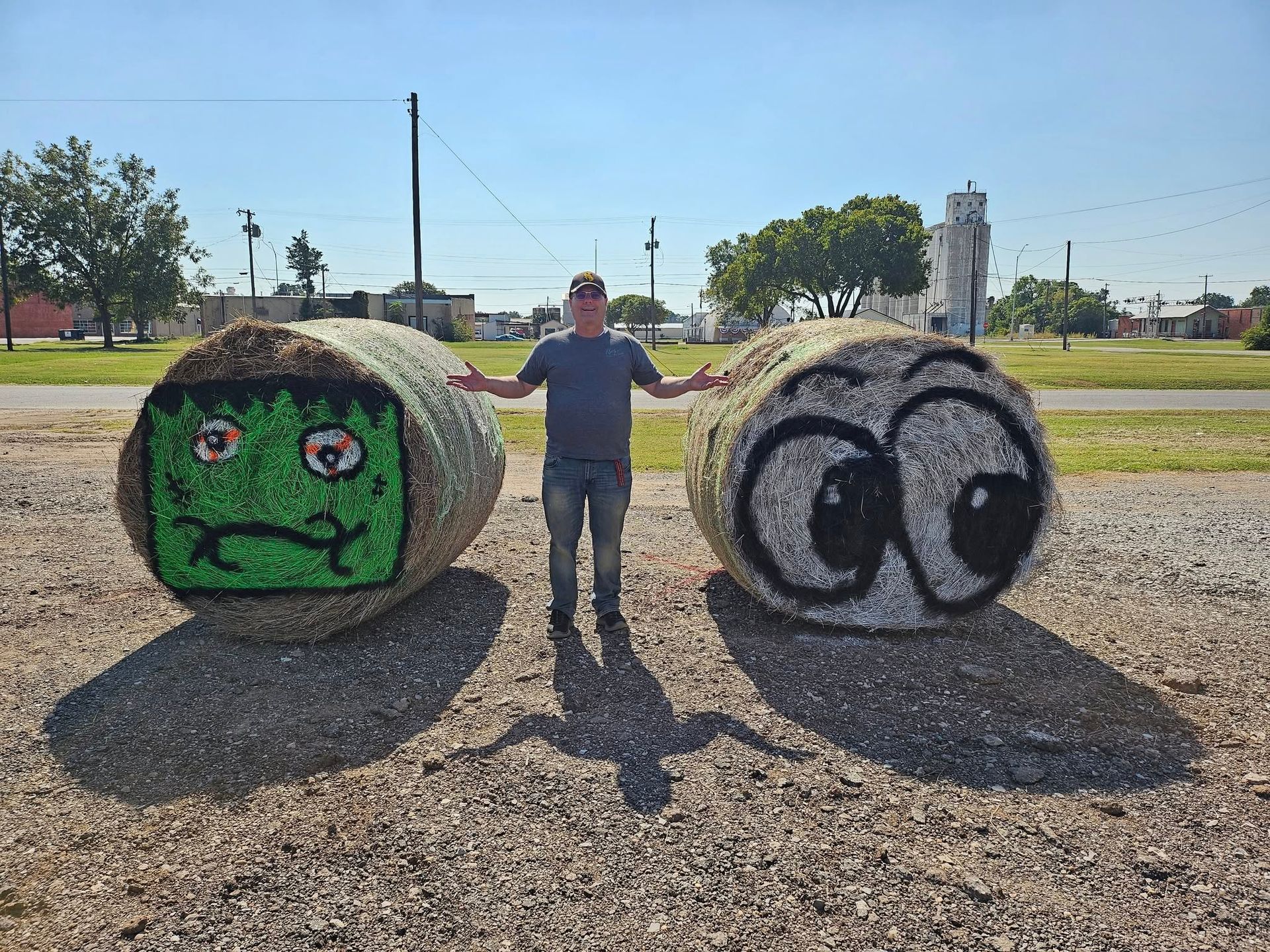 Man stands between two hay bales decorated as cartoon faces in a gravel lot on a sunny day.