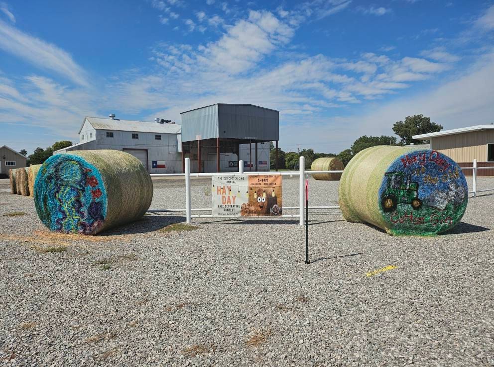Two decorated hay bales, with a sign, in front of buildings under a blue sky.