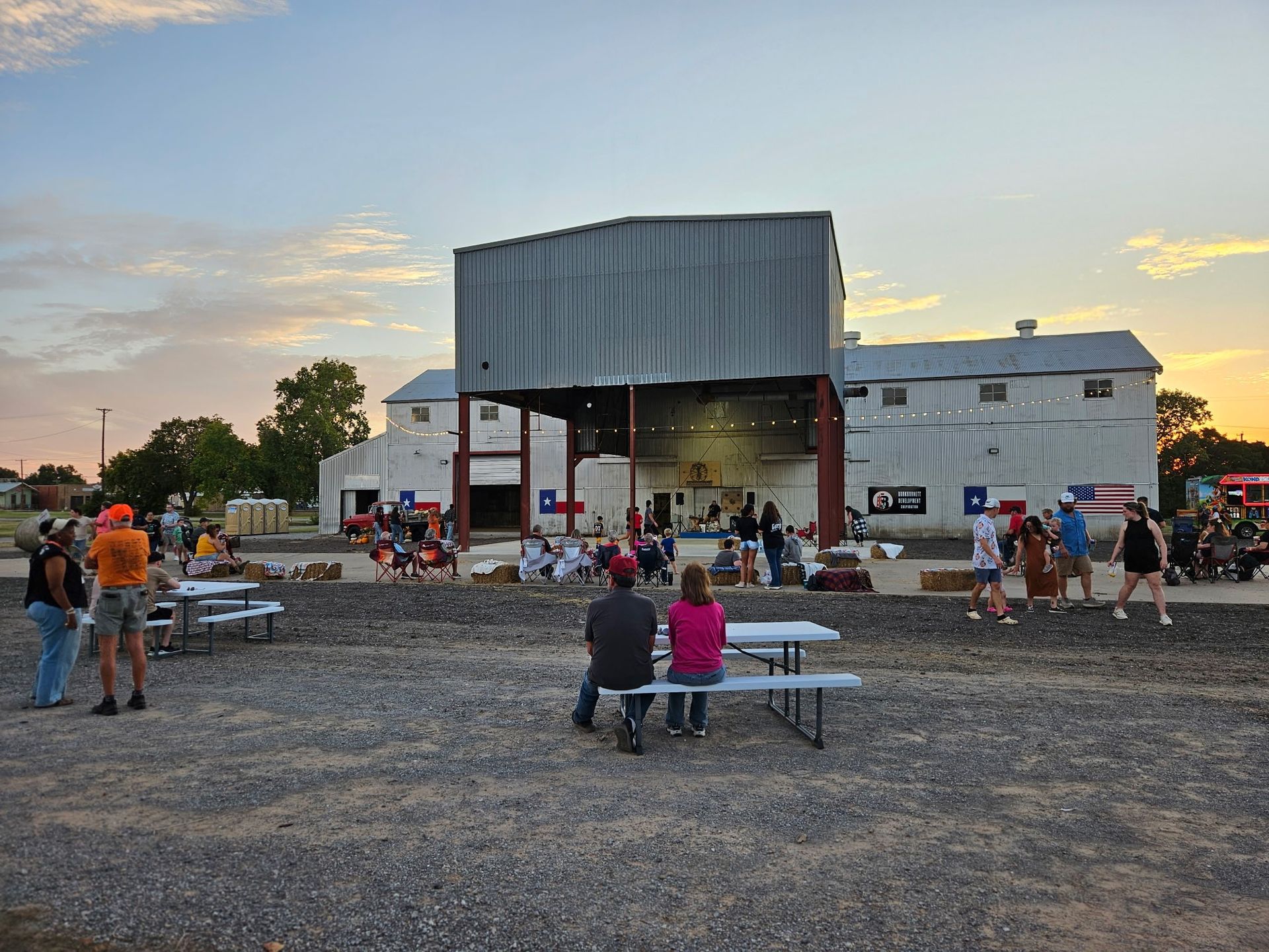 People at an outdoor event with picnic tables, a large white building, and a sunset in the background.
