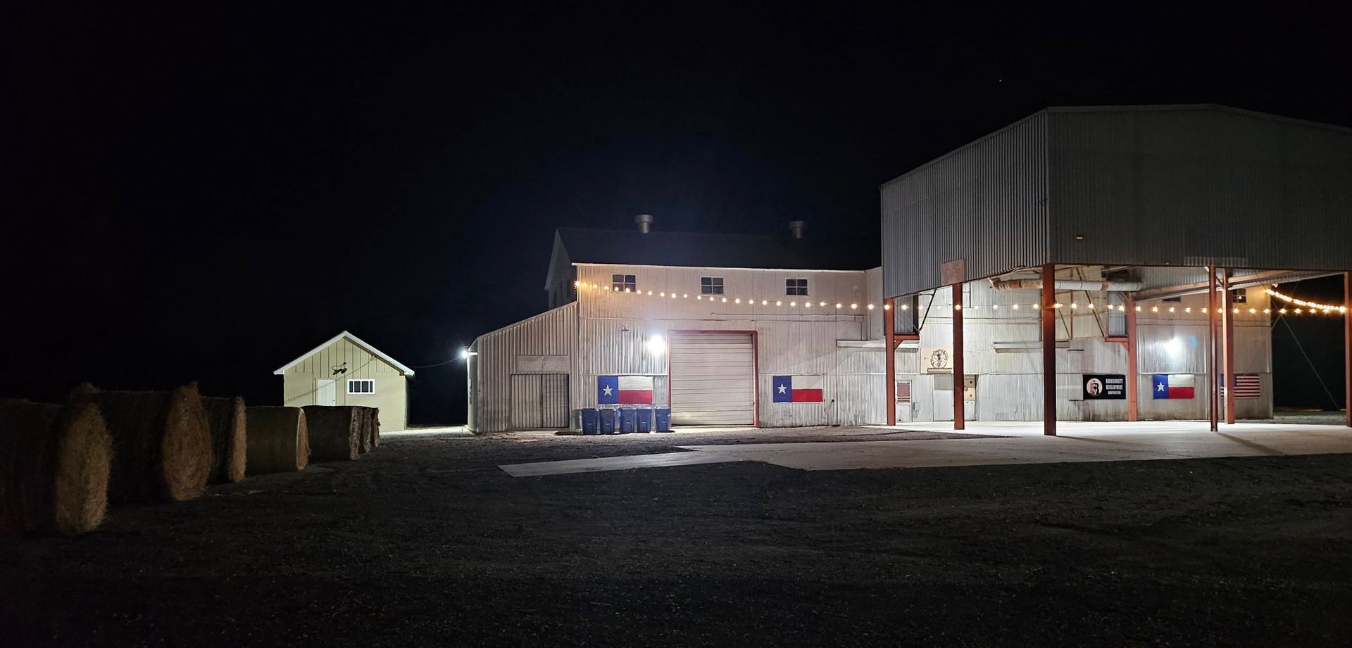 Buildings at night, illuminated by lights. Flags on the wall, hay bales in foreground.