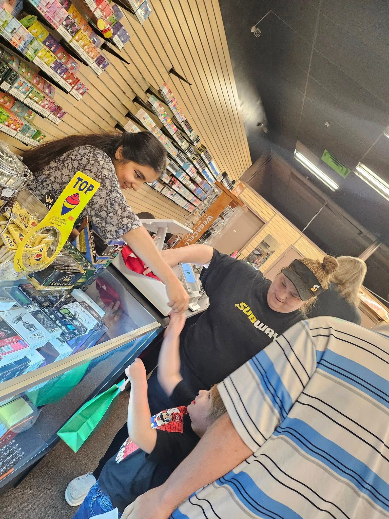 People in a store, reaching for items. One adult is helping a child. Shelves and various products are visible.